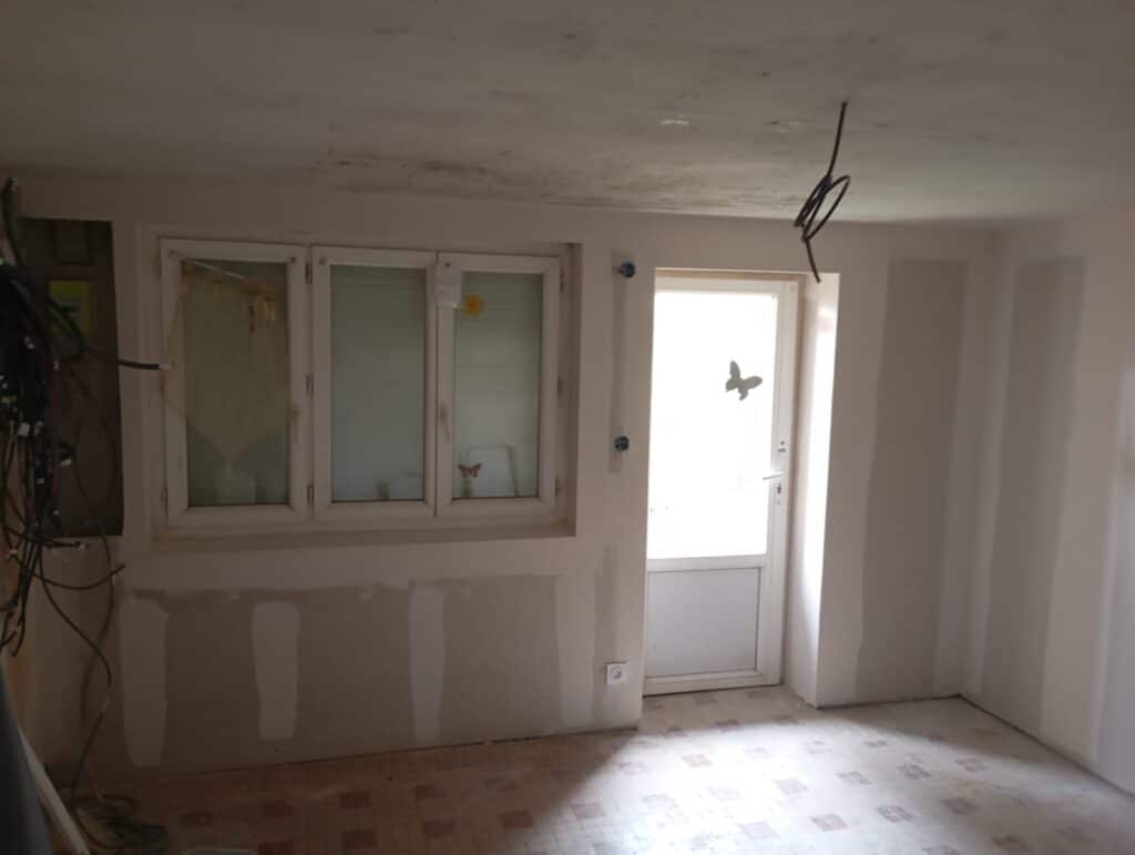 Pièce en travaux : fenêtres blanches, porte vitrée, plaques de plâtre et fils électriques apparents en rénovation.